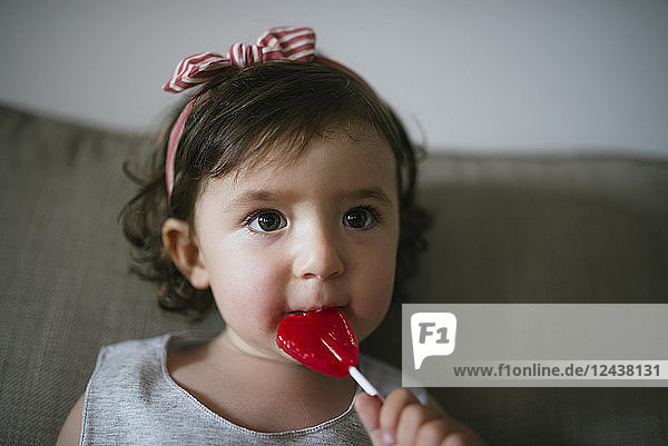 Cute baby girl eating a heart shaped lollipop at home