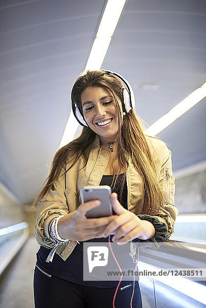 Portrait of smiling young woman with headphones standing on escalator looking at cell phone