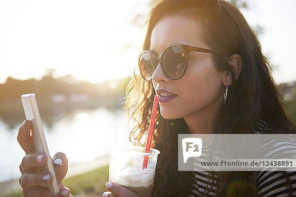 Stylish young woman with cell phone and takeaway drink outdoors at sunset