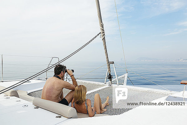 Mature couple sitting on catamaran trampoline  using binoculars