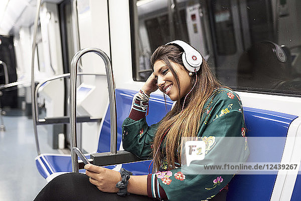 Smiling young woman listening music with headphones and smartphone in underground train