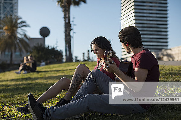 Spain  Barcelona  young man playing guitar for girlfriend sitting on a meadow