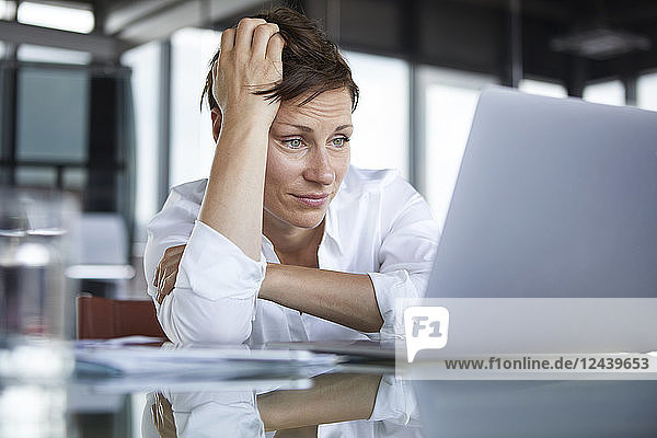 Frustrated businesswoman sitting at glass table in office looking at laptop