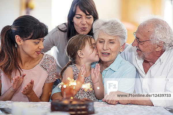 Little girl lwatching sparklers on a birthday cake  sitting on grandmother's lap  with family around