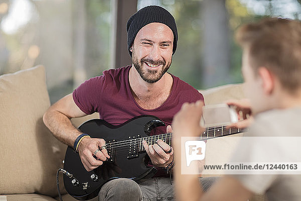 Young man at home sitting on couch playing guitar for teenage boy taking a cell phone picture