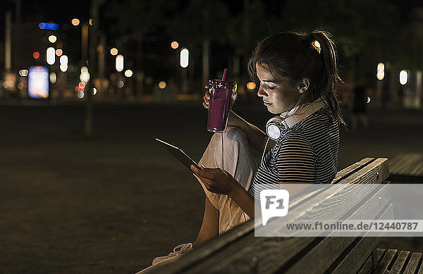 Young woman with smoothie sitting on bench at night using tablet