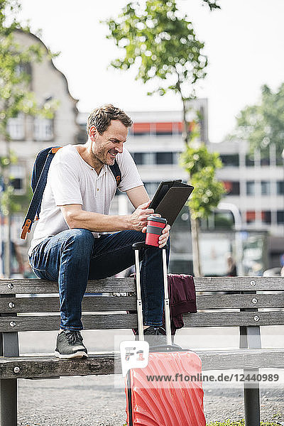 Smiling man with rolling suitcase and takeaway coffee sitting on bench using tablet