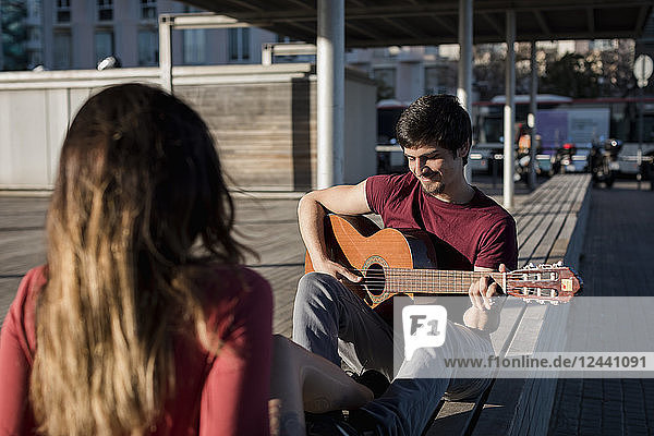 Smiling man sitting on a bench playing guitar for girlfriend