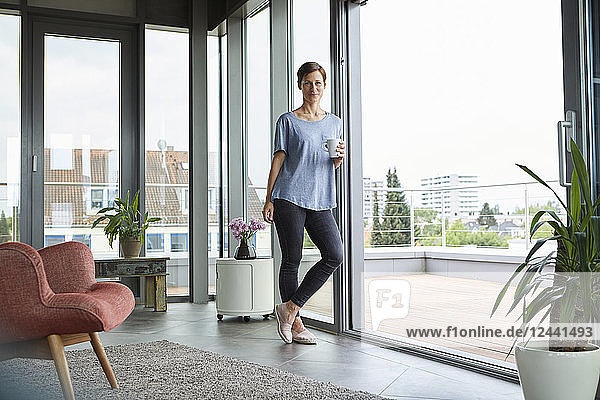 Portrait of woman at home with cup of coffee standing at balcony door