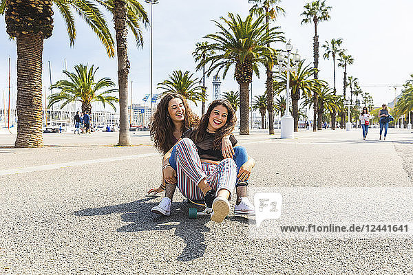 Carefree young woman and teenage girl having fun with a skateboard on a promenade with palms