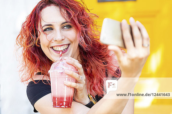Portrait of laughing woman taking selfie with smartphone while drinking smoothie