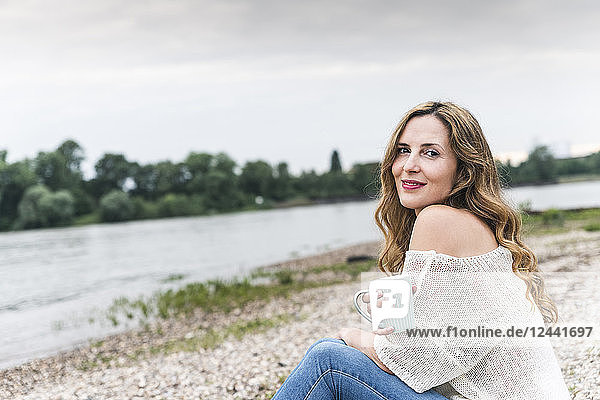 Smiling woman with cup of coffee sitting at the riverside