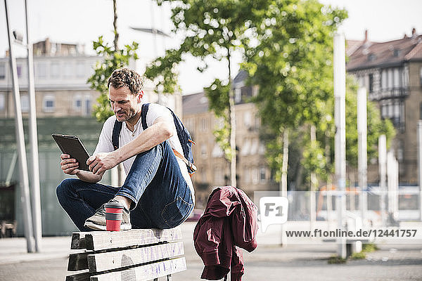 Man with takeaway coffee sitting on bench using tablet