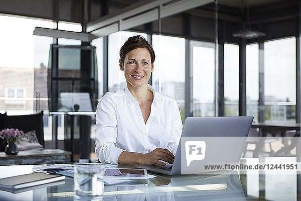 Portrait of smiling businesswoman sitting at glass table in office with laptop