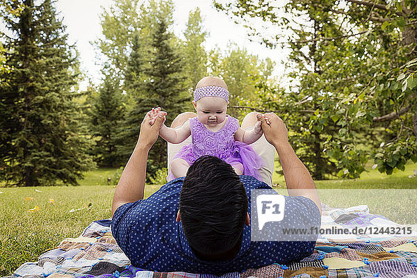 A father playing with his baby daughter while laying on a blanket in a city park in late summer; Edmonton  Alberta  Canada