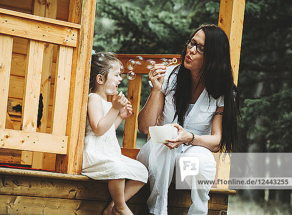 Mother and young daughter blowing bubbles on the steps of a wooden playhouse; Alberta  Canada