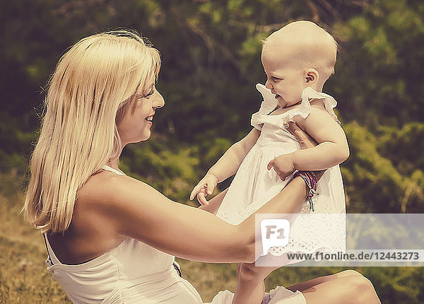 A vintage style image of a beautiful young mother with long blonde hair enjoying quality time with her cute baby daughter sitting on the grass in a city park on a summer day  Edmonton  Alberta  Canada