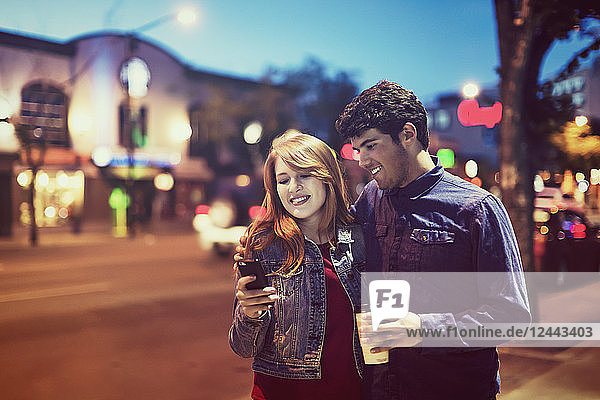 Ein junges Paar,  das in der Abenddämmerung auf einem Bürgersteig in einer beliebten Gegend spazieren geht und dabei auf sein Smartphone schaut,  Edmonton,  Alberta,  Kanada