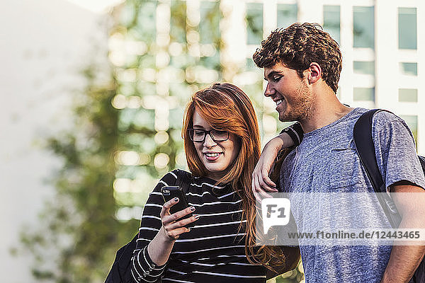 Ein junges Paar,  das zusammen steht und soziale Medien auf einem Smartphone abruft,  während es durch einen Universitätscampus geht,  Edmonton,  Alberta,  Kanada