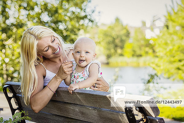 Eine junge Mutter verbringt viel Zeit mit ihrer kleinen Tochter und sitzt auf einer Parkbank neben einem künstlichen See in einem Stadtpark  Edmonton  Alberta  Kanada