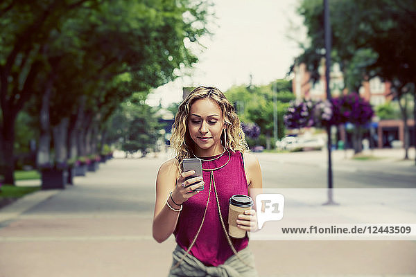 Eine schöne junge Frau  die eine Straße in der Nähe eines Universitätscampus entlangläuft und eine SMS auf ihrem Smartphone schreibt  Edmonton  Alberta  Kanada