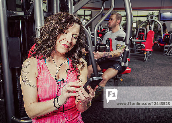 Eine Frau mittleren Alters trainiert in einem Fitnessstudio und hört dabei Musik auf ihrem Smartphone  Spruce Grove  Alberta  Kanada