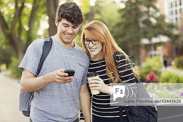 Ein junges Paar  das zusammen steht und soziale Medien auf einem Smartphone abruft  während es durch einen Universitätscampus geht  Edmonton  Alberta  Kanada