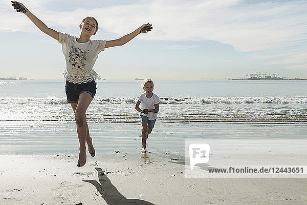 Zwei Schwestern rennen und spielen am Strand  die Älteste vorne nimmt Blickkontakt mit der Kamera auf und springt in die Luft; Long Beach  Kalifornien  Vereinigte Staaten von Amerika