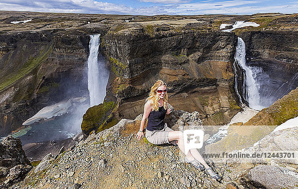 Eine junge Wanderin posiert für ein Porträt am Rande eines atemberaubenden Wasserfalltals,  bekannt als Haifoss,  in Südisland,  Island