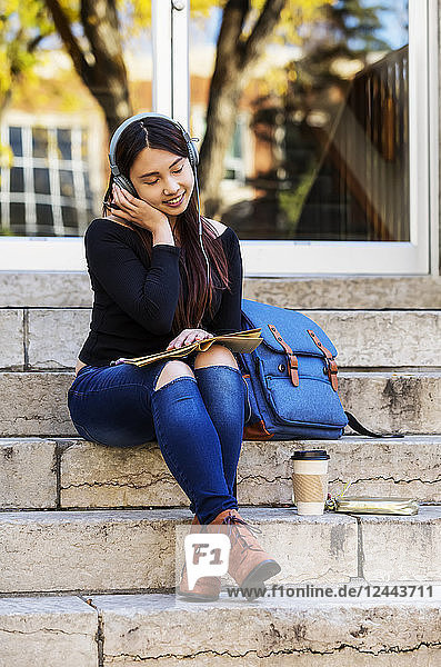 Eine junge chinesische Studentin sitzt auf einer Treppe und hört mit geschlossenen Augen über Kopfhörer Musik  Edmonton  Alberta  Kanada