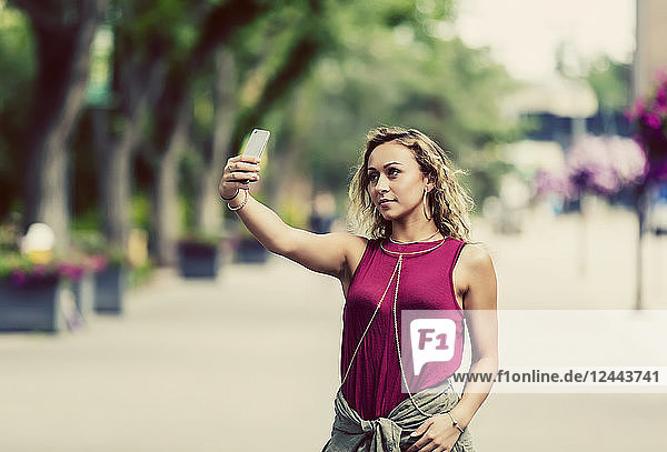 Eine konzeptionelle Aufnahme einer jungen Frau  die ein Selbstporträt mit ihrem Smartphone aufnimmt  während sie allein auf der Straße in der Nähe eines Universitätsgeländes unterwegs ist  Edmonton  Alberta  Kanada