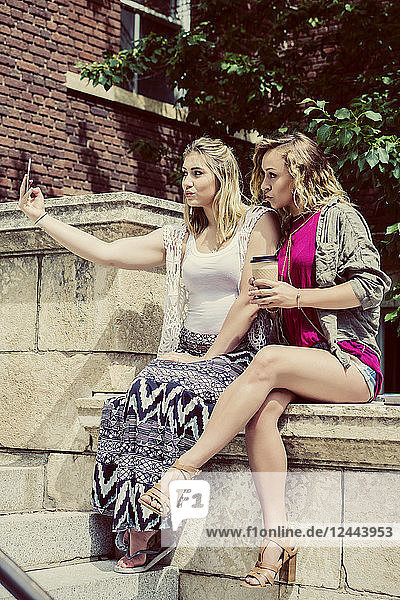 Two female university students sitting together on the campus taking a self-portrait with their smart phone and making silly pouty lips  Edmonton  Alberta  Canada