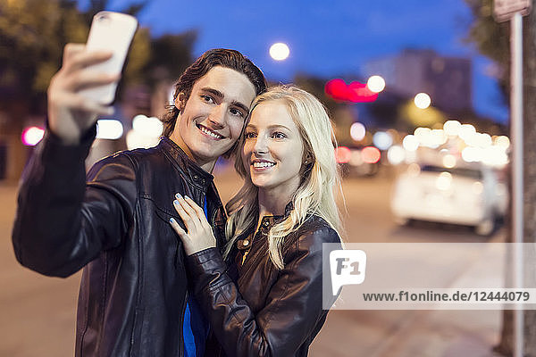 Ein junges Paar macht ein Selbstporträt mit einem Smartphone entlang einer Straße in der Abenddämmerung,  Edmonton,  Alberta,  Kanada