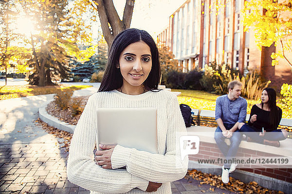Eine junge Universitätsstudentin libanesischer Abstammung posiert mit ihrem Laptop in der Hand  während sich ihre Freunde im Hintergrund unterhalten  Edmonton  Alberta  Kanada
