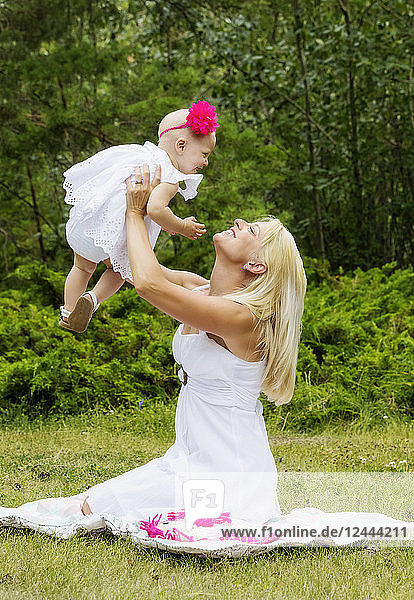 Eine hübsche junge Mutter mit langen blonden Haaren genießt die Zeit mit ihrer süßen kleinen Tochter und wirft sie in die Luft  während sie an einem Sommertag im Gras eines Stadtparks sitzt  Edmonton  Alberta  Kanada