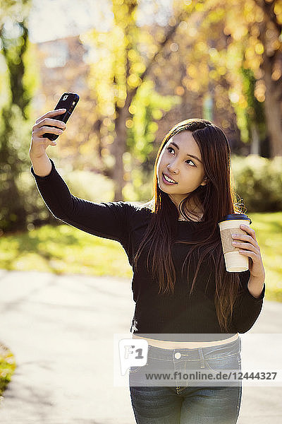 Eine junge internationale Universitätsstudentin hält eine Kaffeetasse in der Hand und posiert für ein Selbstporträt mit ihrem Smartphone  Edmonton  Alberta  Kanada