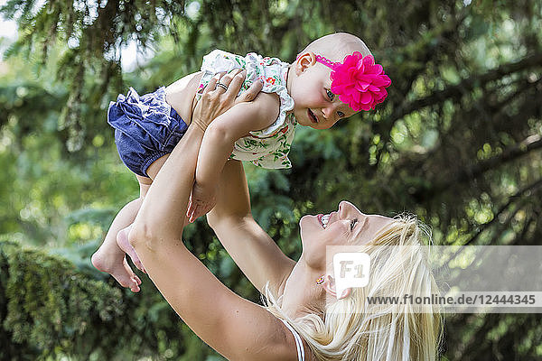 A beautiful young mother with long blonde hair enjoying quality time with her cute baby daughter and tossing her in the air in a city park on a summer day  Edmonton  Alberta  Canada