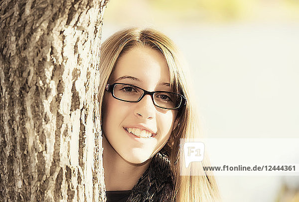 Portrait of a young girl with eyeglasses peering out from behind a tree in autumn  Edmonton  Alberta  Canada