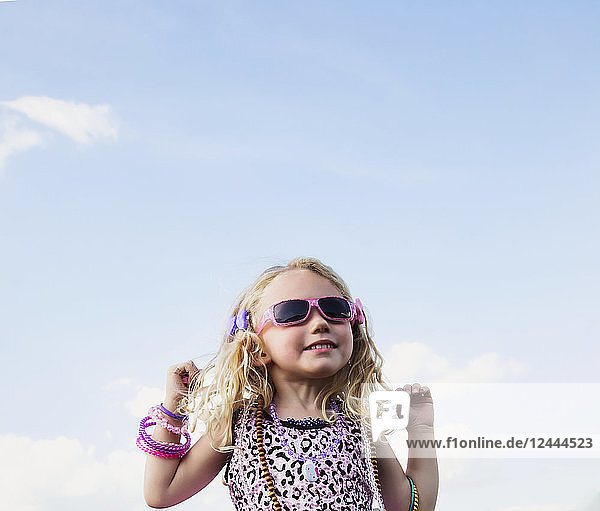 Ein junges Mädchen mit blondem  lockigem Haar  Sonnenbrille und Schmuck  das vor einem blauen Himmel mit Wolken steht und niedliche Grimassen schneidet; Spruce Grove  Alberta  Kanada