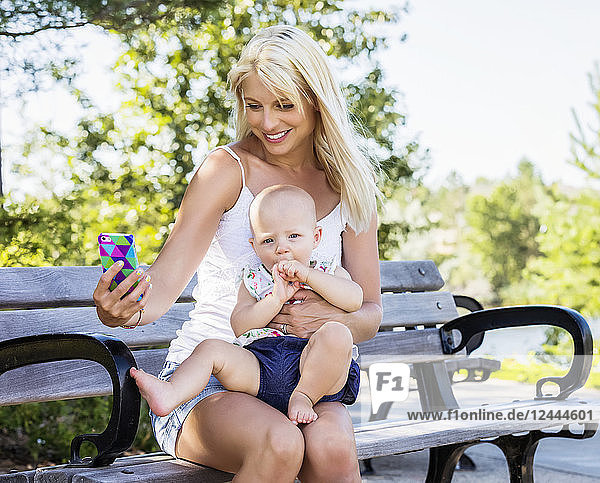 A beautiful young mom with her baby daughter sitting on a park bench and trying to take a self-portrait while spending quality time together outdoors on a warm summer day  Edmonton  Alberta  Canada