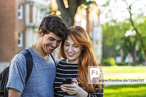 Ein junges Paar,  das zusammen steht und soziale Medien auf einem Smartphone abruft,  während es durch einen Universitätscampus geht,  Edmonton,  Alberta,  Kanada