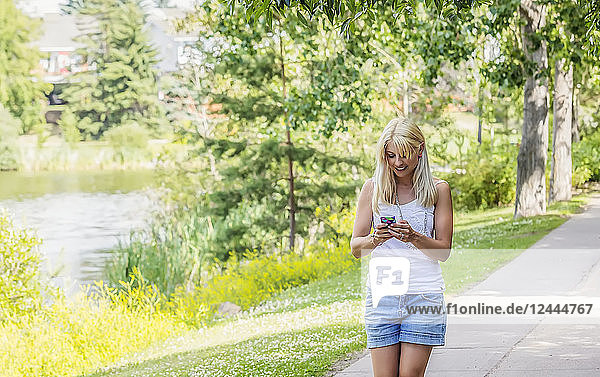 Eine schöne junge Frau  die an einem warmen sonnigen Tag durch einen Park an einem See spaziert und eine SMS auf ihrem Smartphone schreibt  Edmonton  Alberta  Kanada