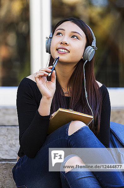 A young Chinese female university student sits on the steps listening to music using headphones and looks up while daydreaming and holding a pen top to her chin  Edmonton  Alberta  Canada