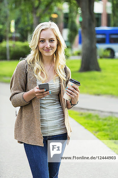 Eine schöne junge Frau mit langen blonden Haaren hält eine Kaffeetasse und schreibt eine SMS auf ihrem Smartphone,  während sie über einen Universitätscampus geht,  Edmonton,  Alberta,  Kanada
