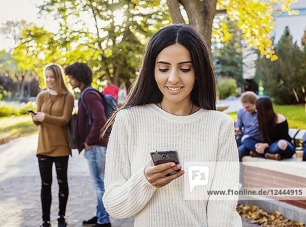 Eine junge Studentin libanesischer Herkunft überprüft ihr Smartphone  während sich ihre Freunde im Hintergrund auf dem Universitätscampus unterhalten  Edmonton  Alberta  Kanada