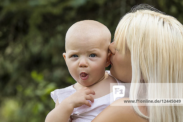 Eine hübsche junge Mutter kuschelt mit ihrer kleinen Tochter  die ein lustiges Gesicht macht  während sie an einem warmen Sommertag in einem Park Zeit miteinander verbringt  Edmonton  Alberta  Kanada