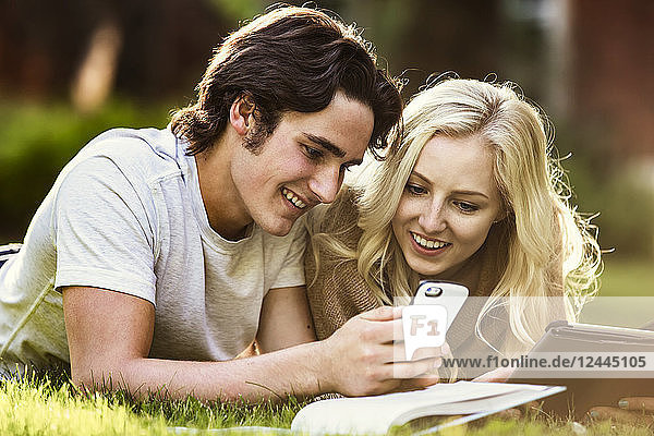 Ein junges Paar studiert im Freien auf dem Rasen des Universitätscampus mit einem Lehrbuch und einem Tablet und überprüft soziale Medien auf einem Smartphone  Edmonton  Alberta  Kanada
