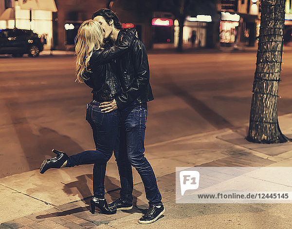 Ein junges Paar in schwarzen Lederjacken,  die Frau hebt ein Bein,  während sie nachts auf einem Gehweg stehen und sich küssen,  Edmonton,  Alberta,  Kanada