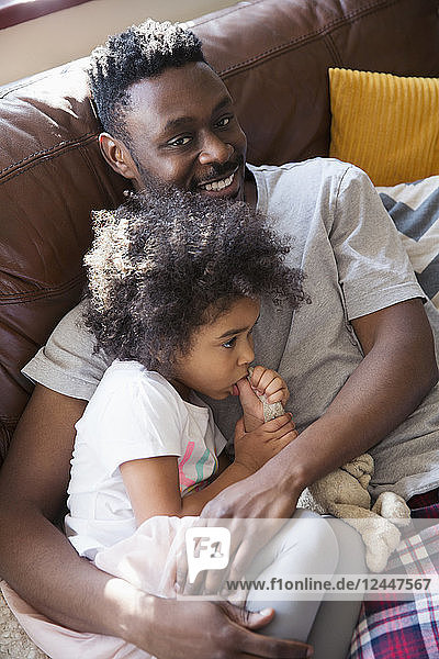 Porträt liebevoller Vater  der mit seiner kleinen Tochter auf dem Sofa kuschelt und am Daumen lutscht
