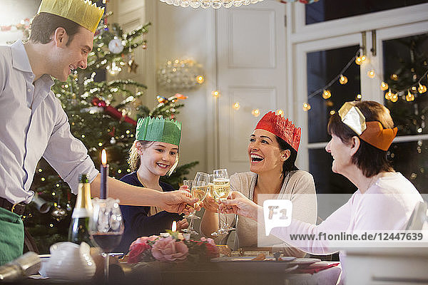 Mehrgenerationenfamilie in Papierkronen stößt beim Weihnachtsessen bei Kerzenschein mit Champagnerflöten an